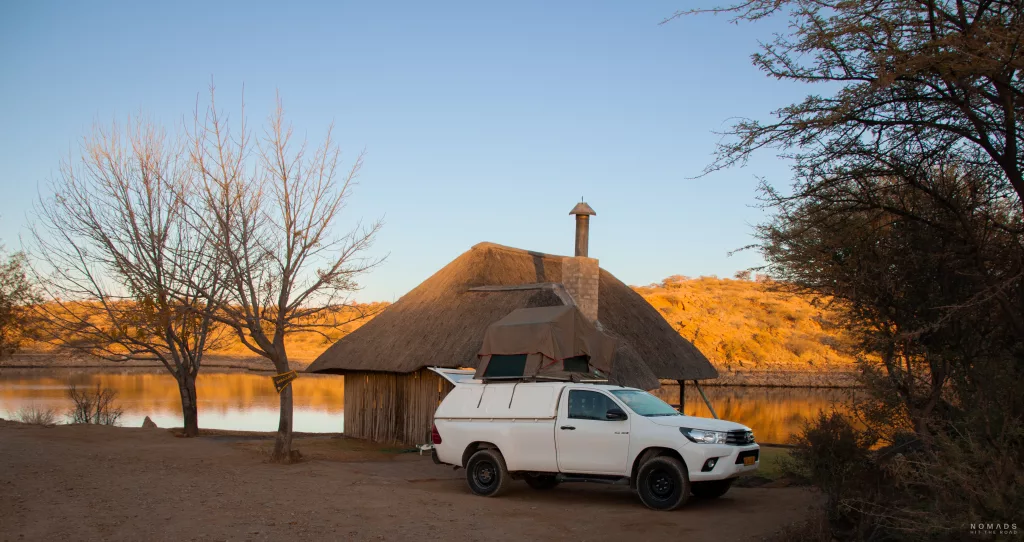 Weißer Pickup mit aufgestelltem Dachzelt steht auf einem Campingplatz mit Hütte am Lake Oanob in Namibia. Im Hintergrund der perfekte Sonnenaufgang.