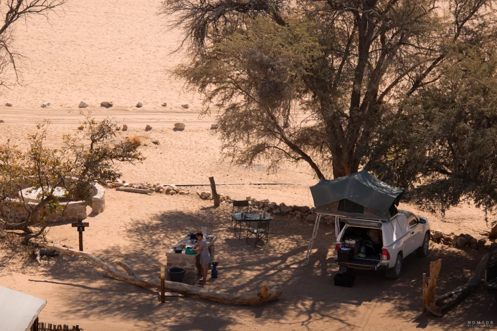 Campingplatz von oben mit Auto inkl. Dachzelt auf der rechten Seite, in der Mitte Küchenzeil mit Frau und links Feuerstelle