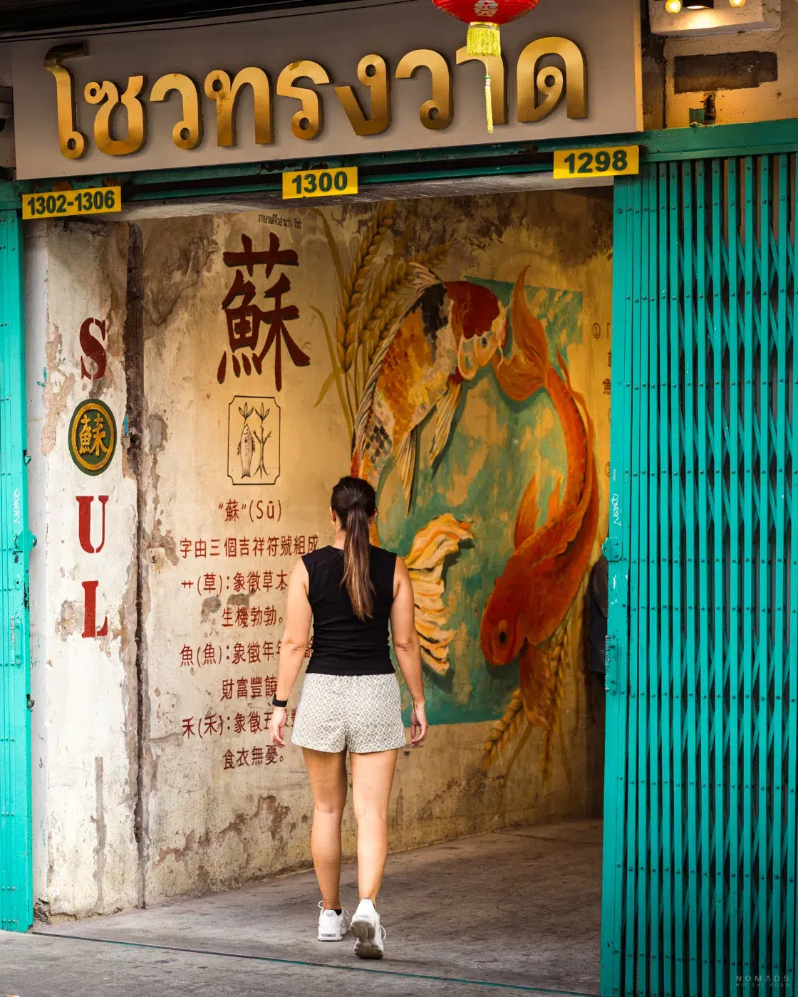 Rückansicht einer jungen Frau, die durch ein geöffnetes, türkisfarbenes Metalltor in den Eingangsbereich des „Soul Songwat“ in Bangkok geht. An der verwaschenen Wand im Inneren ist ein großes Kunstwerk mit zwei Koi-Fischen und chinesischen Schriftzeichen zu sehen.