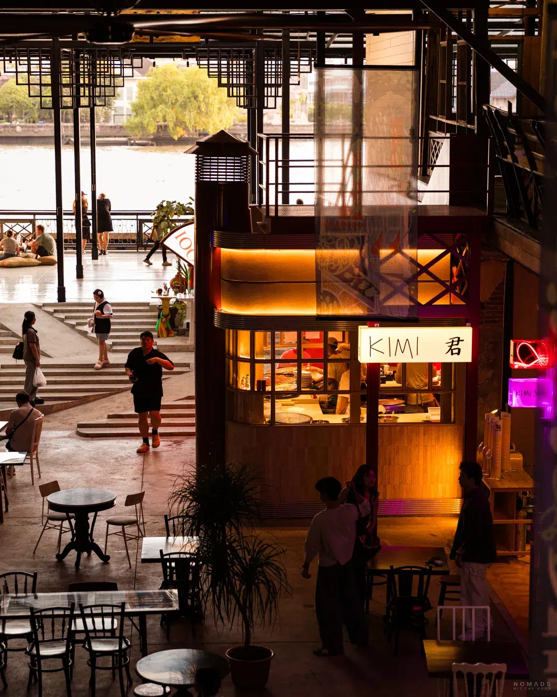 Ein moderner, halboffener Community Space an der Song Wat Road in Bangkok am Chao Phraya Fluss in der Abenddämmerung. Zu sehen sind Tische, ein hell beleuchteter Imbissstand namens „KIMI“ und Besucher, die auf einer Terrasse am Wasser sitzen.