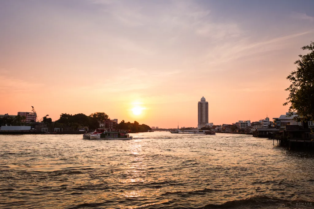 Sonnenuntergang über dem Chao Phraya Fluss in Bangkok. Die Sonne steht tief am orange-rosa gefärbten Himmel, während ein Passagierboot auf dem Wasser fährt. Am gegenüberliegenden Ufer sind Gebäude, ein markantes Hochhaus mit einer Kuppel und Vegetation zu sehen.