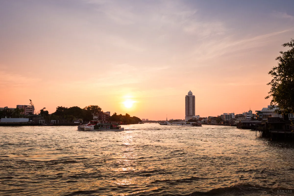 Sonnenuntergang über dem Chao Phraya Fluss in Bangkok. Die Sonne steht tief am orange-rosa gefärbten Himmel, während ein Passagierboot auf dem Wasser fährt. Am gegenüberliegenden Ufer sind Gebäude, ein markantes Hochhaus mit einer Kuppel und Vegetation zu sehen.