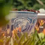 Nahaufnahme eines kunstvoll verzierten blauen Keramiktopfes in Ancient City, Thailand, umgeben von Pflanzen und Blumen.