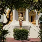 Statue einer thailändischen Figur in betender Pose vor einem kunstvoll verzierten Tempel in Ancient City, Thailand, umgeben von grüner Vegetation.