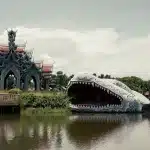 Traditioneller thailändischer Tempel mit kunstvollen Dächern neben einer riesigen weißen Drachenfigur am Ufer eines Sees in Ancient City, Thailand.