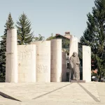 Denkmal in Bansko, Bulgarien, mit einer Statue und Steinsäulen im Hintergrund, umgeben von hohen Bäumen und traditioneller Architektur.