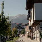 Ruhige Straße in Bansko mit Blick auf das Pirin-Gebirge im Hintergrund, traditionelle Häuser und eine Frau, die den Gehweg entlanggeht.