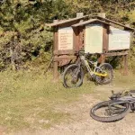 Zwei E-Mountainbikes stehen vor einer Wander- und Trailkarte im Pirin-Nationalpark, umgeben von Natur und Bäumen.