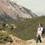 Wanderin mit Rucksack genießt die Aussicht im Pirin-Gebirge, umgeben von bewaldeten Hängen und steilen Felswänden unter blauem Himmel.