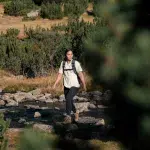 Wanderin überquert einen kleinen Bergbach im Pirin-Gebirge, umgeben von alpiner Vegetation und Felsen an einem sonnigen Tag.