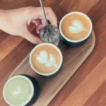 Drei Tassen Cappuccino mit Milchschaum-Herzen, auf einem Holztablett serviert, daneben ein Glas Wasser und ein Löffel in einer Hand.