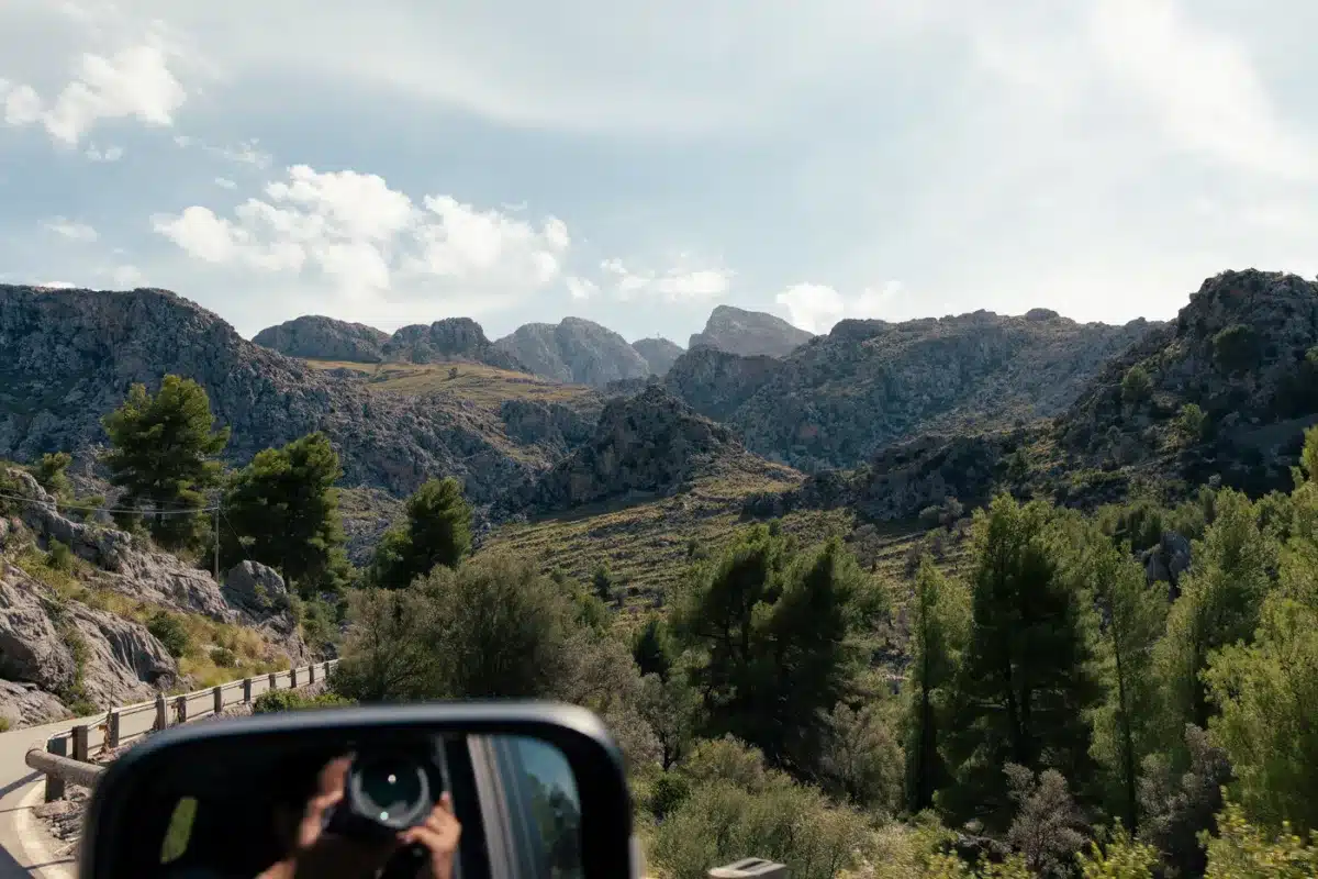 Blick aus dem Autofenster auf die MA10 Mollarcas, mit Bergpanorama und Pinienbäumen entlang der Straße, perfekt für einen Roadtrip entlang Mallorcas Westküste.
