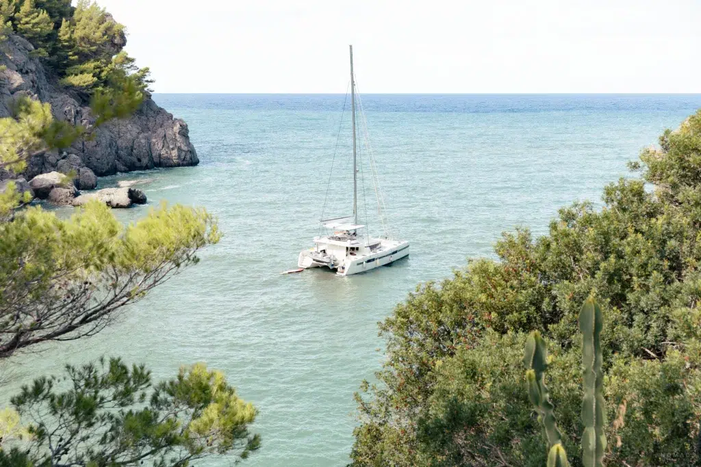 Katamaran vor dem kleinen Hafen von Sa Calobra auf Mallorca, umgeben von ruhigem, blau-grünem Wasser und eingerahmt von Felsen und Pinien.