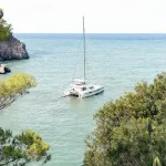 Katamaran vor dem kleinen Hafen von Sa Calobra auf Mallorca, umgeben von ruhigem, blau-grünem Wasser und eingerahmt von Felsen und Pinien.