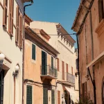 Eine sonnige Gasse in Santanyi auf Mallorca mit traditionellen mediterranen Gebäuden, grünen Fensterläden und Holzläden, unter einem strahlend blauen Himmel.