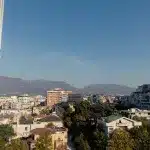 Panoramaansicht von Tirana: Eine weitläufige Stadtansicht mit modernen und traditionellen Gebäuden im Vordergrund, vor der Kulisse beeindruckender Berge und klarem, blauem Himmel.