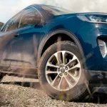 Ein blauer Ford Kuga aus der Nahperspektive, mit staubigen Reifen, die seine Offroad-Tauglichkeit betonen, vor einer malerischen Berglandschaft.