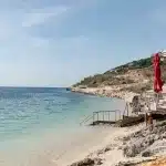 Sandstrand mit klarem, türkisfarbenem Wasser und roten Sonnenschirmen – ein ruhiger Küstenabschnitt in Albanien.