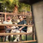 Zwei Personen entspannen in einem gemütlichen Restaurant mit Blick auf Palmen und Holzmöbel, genießen die warme Atmosphäre.