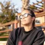 Frau mit Sonnenbrille genießt die Abendsonne in einer entspannten Outdoor-Location, umgeben von Palmen und warmem Licht.