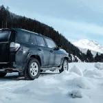 Schwarzes Auto steht am verschneiten Straßenrand, während auf der anderen Seite ein tannenwald steht