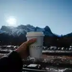 Person hält einen Kaffeebecher von Eclipse aus dem Elevation Place in Canmore in der Hand. Im Hintergrund wunderschöne Berge zu sehen.