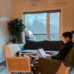 Frau sitz auf einem Stuhl mit ihrem Laptop in einer Wohnung. Durch die Fenster nach draußen zeigen sich die Berge, die Canmore umgeben