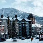 Blick auf Whistler Village mit alpiner Architektur, einer großen Uhr und schneebedeckten Bergen im Hintergrund, während Fußgänger und Autos die Straßen beleben.