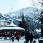 Winterliche Szenerie in Whistler Village mit verschneiten Gebäuden, einer überdachten Sitzgelegenheit und Menschen mit Snowboardausrüstung vor dem Hintergrund schneebedeckter Berge.