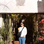 Frau mit Sonnenbrille und Orangensaft steht im Eingang eines charmanten Cafés mit Pflanzen und Boho-Flair – entspannte Stimmung am Morgen.
