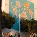 Wandgemälde mit Porträts und Naturmotiven im Abendlicht an einer Hauswand in La Paz – darunter Straßenleben mit Verkaufsstand und Passanten.