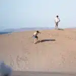 Zwei junge Männer beim Sandboarden auf den Dünen von El Mogote – Spaß und Sport mit Meerblick.