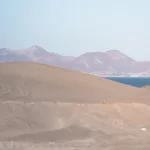 Weiche Sanddünen treffen auf das blaue Meer – im Hintergrund die Küstenlinie von La Paz mit Blick auf die Berge.