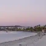 Strandpromenade von La Paz im sanften Abendlicht mit Blick auf den Stadtstrand, Palmen und vorbeiziehenden Jetski-Fahrern vor rosa Himmel.