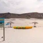Gestapelte gelbe und grüne Kajaks auf dem Strand von Balandra mit Warnschild vor Rochen und Palapa-Sonnenschirmen im Hintergrund.
