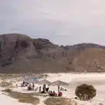 Menschen entspannen sich unter Palapa-Sonnenschirmen am weißen Sandstrand von Balandra, umgeben von trockenen Bergen unter bewölktem Himmel.