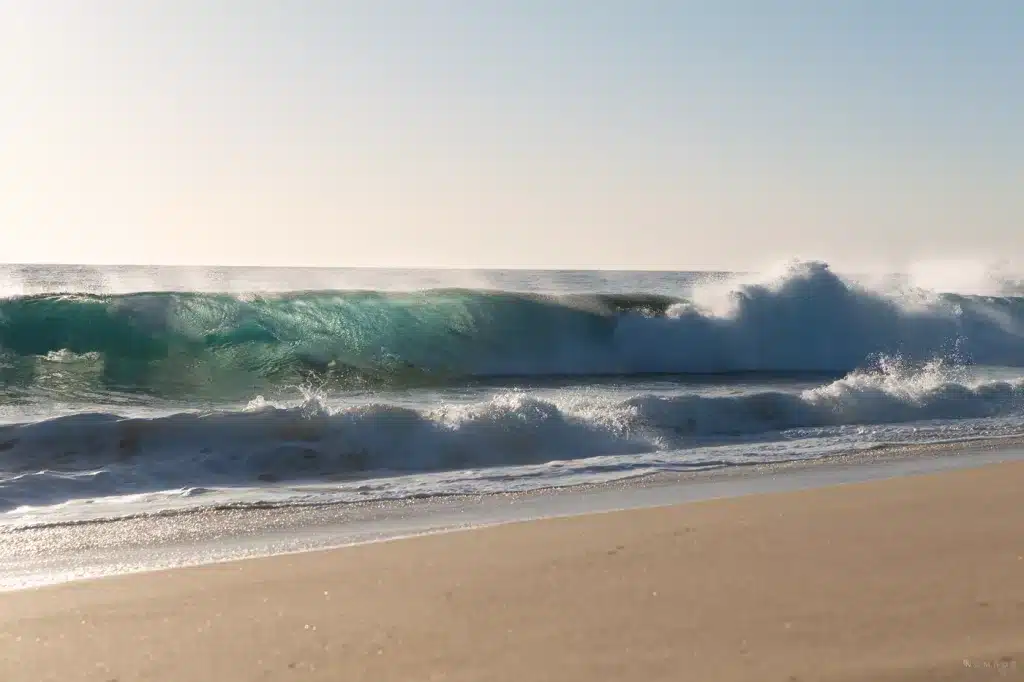 Eine kräftige Welle bricht mit voller Kraft an einem naturbelassenen Strand in der Nähe von Todos Santos, Mexiko – türkis schimmerndes Wasser im Licht der untergehenden Sonne.