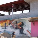 Street Art eines Surfers auf einer Hauswand, während davor ein Mann auf der Baustelle arbeitet