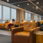 Menschen sitzen entspannt in modernen Loungesesseln mit Blick auf die Rollbahn des Flughafens.
