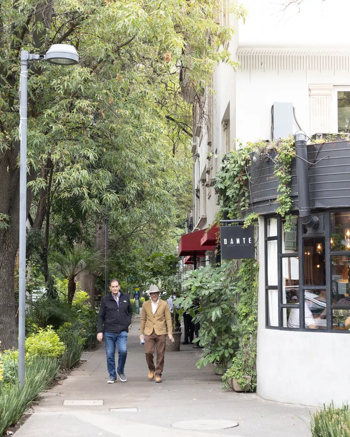 Zwei Männer laufen auf einem Bürgersteig, der gesäumt ist von Restaurants und Bäumen