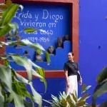 Der Innenhof der Casa Azul, dem ehemaligen Wohnhaus von Frida Kahlo, mit einem leuchtend blauen Gebäude und üppiger grüner Vegetation. Ein großer Baum mit einem dicken Stamm dominiert die Mitte des Hofes, und im Hintergrund sind weitere Teile des Hauses zu sehen.