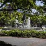 Die Fuente de Cibeles, ein berühmter Brunnen in Mexiko-Stadt, der von einem großen, runden Platz umgeben ist. Der Brunnen zeigt Statuen und Wasserspiele, umgeben von Bäumen und Gebäuden im Hintergrund. Am oberen Rand des Bildes ist ein klarer, heller Himmel zu sehen.