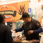 Eine Ansicht eines Marktstandes auf dem Tianguis los Martes, der eine breite Palette an frischem Obst und Gemüse anbietet, darunter Bananen, Papayas und grüne Blätter. Die Produkte sind auf einem Tisch ausgebreitet, und im Hintergrund sind weitere Marktstände und Menschen zu sehen, die durch den Markt schlendern.