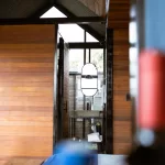 Blick vom Bett im Boato Hotel in Guatapé, Kolumbien, durch den offenen Raum auf das moderne Badezimmer mit Holz- und Glaselementen, im Vordergrund unscharfe Flasche.
