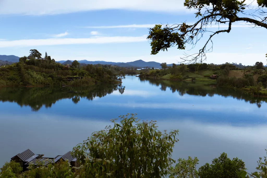 Panoramablick auf den Stausee von Guatapé, Kolumbien, mit spiegelglattem Wasser, grünen Inseln und Bergen im Hintergrund, unten im Bild moderne Hütten des Boato Hotels.