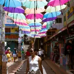 Touristin geht durch farbenfrohe Gasse in Guatapé, Kolumbien, unter einer Installation aus bunten Regenschirmen