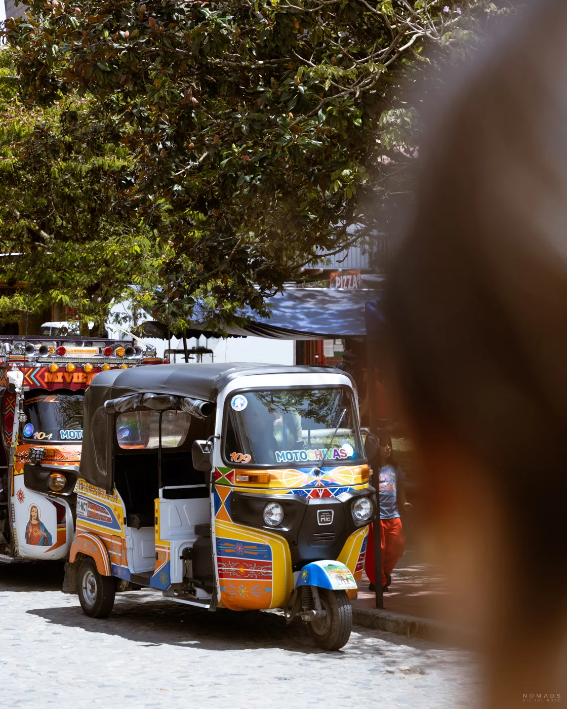 Bunt bemaltes TukTuk in Guatapé, Kolumbien, vor Bäumen und Straßenständen – typisches Transportmittel für Touristen