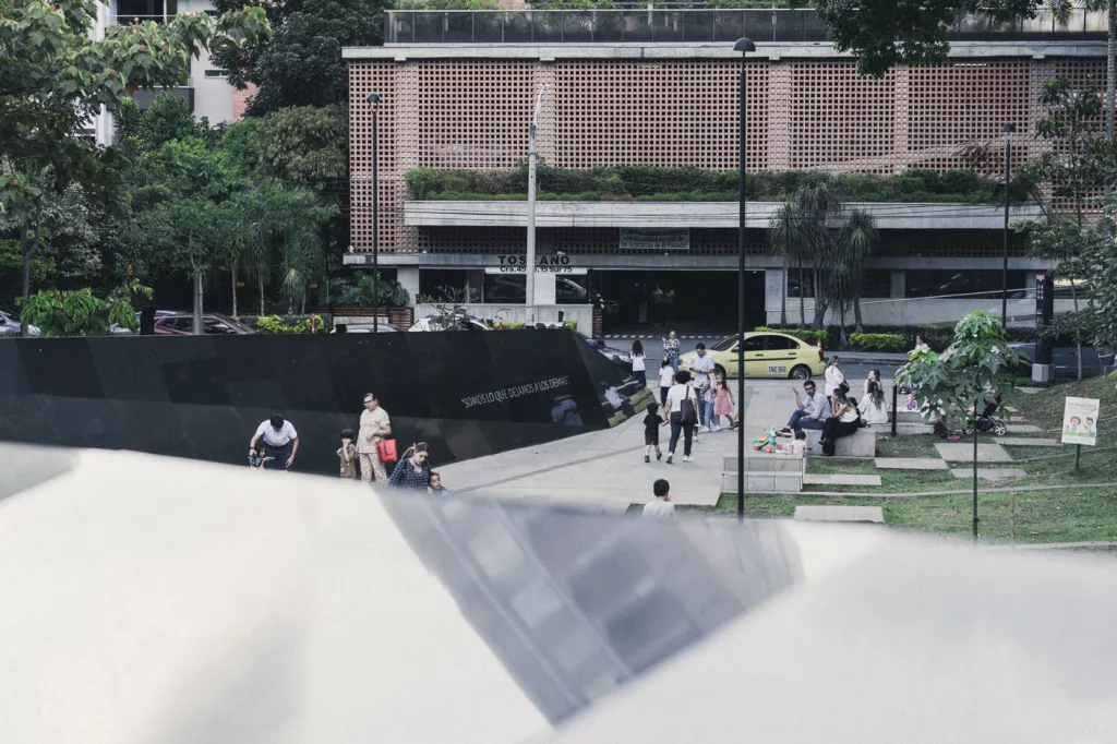 Gedenkpark Inflexión in Medellín mit schwarzer Gedenkmauer, auf der Inschriften zu sehen sind. Menschen sitzen und spazieren durch den Park, der an die Opfer der Gewalt des Medellín-Kartells erinnert.
