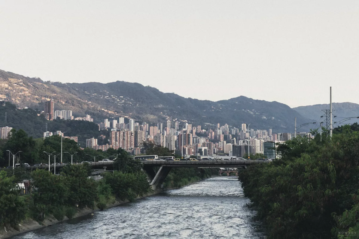 Blick über Medellín mit dem Río Medellín im Vordergrund, Brücke mit Verkehr und dicht bebauten Hochhäusern vor der Kulisse der Anden.