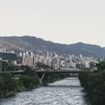 Blick über Medellín mit dem Río Medellín im Vordergrund, Brücke mit Verkehr und dicht bebauten Hochhäusern vor der Kulisse der Anden.